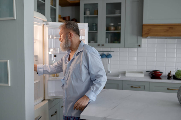 Senior man hunger looking inside open fridge on kitchen searching for snack. Old male suffering from eating disorder, unhealthy food addiction - Zdjęcie, obraz