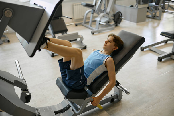 Προεφηβικό αγόρι προπόνηση πόδια σε αθλητικό εξοπλισμό στο γυμναστήριο. Νεαρός αθλητής σπρώχνει πλάκα με τα πόδια για να είναι ισχυρή και την υγεία - Φωτογραφία, εικόνα