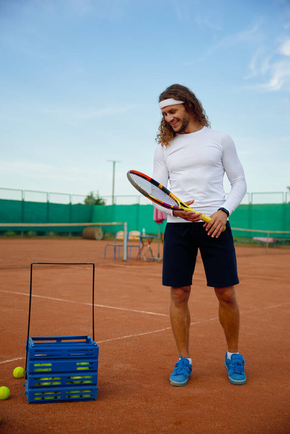 Νεαρός ικανοποιημένος παίκτης τένις μαζεύοντας διάσπαρτες μπάλες στο γήπεδο μετά την προπόνηση του παιχνιδιού. Επαγγελματική έννοια ή έννοια χόμπι αναψυχής - Φωτογραφία, εικόνα