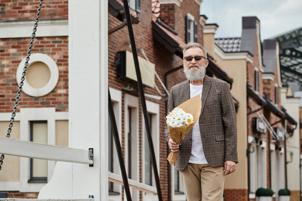 uomo anziano con barba e occhiali da sole in possesso di mazzo di fiori, in piedi sulla strada urbana, elegante - Foto, immagini
