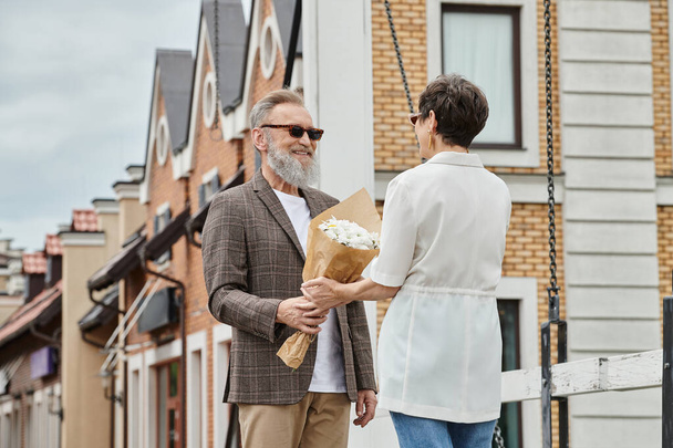 homme âgé heureux avec barbe et lunettes de soleil à la mode donnant bouquet à la femme sur la rue, date, romance - Photo, image