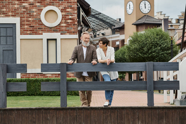 glückliches älteres Paar, Frau schaut Mann an, steht am Zaun, urbane Kulisse, alternde Bevölkerung - Foto, Bild
