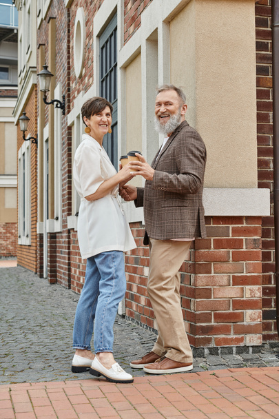 счастливая пожилая пара держит кофе, чтобы пойти, здание, пожилой мужчина и женщина смотрят в камеру, свидание - Фото, изображение