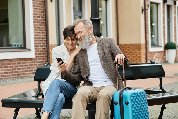 elderly couple, happy senior man holding smartphone, sitting with woman on bench, luggage, gadget - Photo, Image