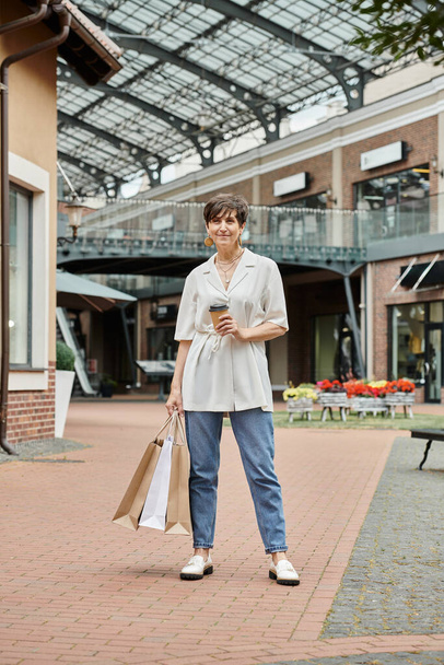 starší žena s krátkými vlasy drží nákupní tašky a kávu jít do blízkosti outlet venku, starší - Fotografie, Obrázek
