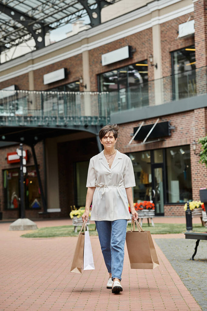 tevreden senior vrouw wandelen met boodschappentassen op stedelijke straat, detailhandel, ouderen in de stad, buiten - Foto, afbeelding