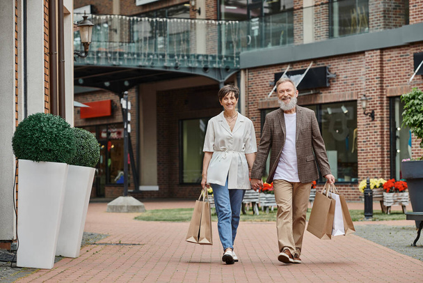 szczęśliwy starsza para mając zakupy, starszy mężczyzna i kobieta trzymając się za ręce i spacerując po ulicy - Zdjęcie, obraz