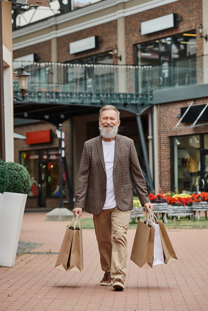 χαρούμενος γενειοφόρος άνδρας με τα πόδια με τσάντες ψώνια, τη ζωή των τελειόφοιτων, αστικού δρόμου, θετική, κομψή στολή - Φωτογραφία, εικόνα