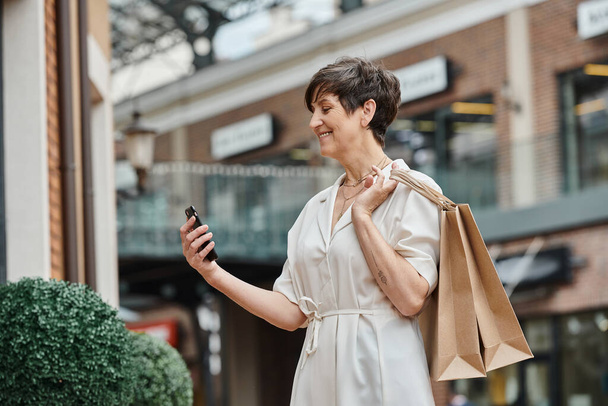 heureuse femme âgée en utilisant smartphone, tenant des sacs à provisions et debout près du centre commercial extérieur - Photo, image