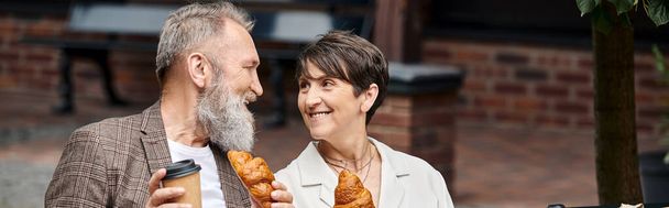 happy elderly couple holding croissants and coffee to go, paper cup, outdoors, banner horizontal - Photo, Image