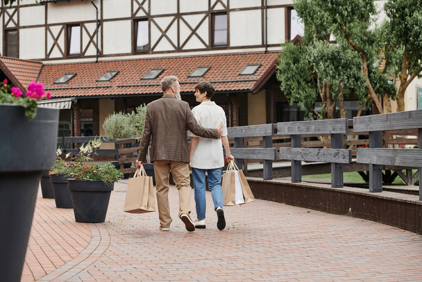 Rückansicht eines älteren Paares, das mit Einkaufstaschen spaziert, älterer Mann umarmt Frau, urbaner Lebensstil - Foto, Bild