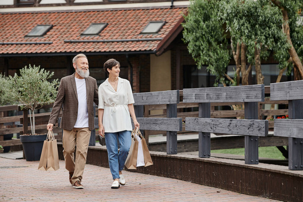θετικό ζευγάρι ηλικιωμένων που περπατά με τσάντες για ψώνια, ηλικιωμένος άντρας που αγκαλιάζει γυναίκα, περπατά μαζί - Φωτογραφία, εικόνα