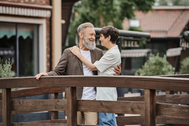 ρομαντικό ζευγάρι ηλικιωμένων που στέκονται μαζί σε ξύλινη γέφυρα και αγκαλιάζονται, ηλικιωμένος έρωτας, δέσιμο - Φωτογραφία, εικόνα