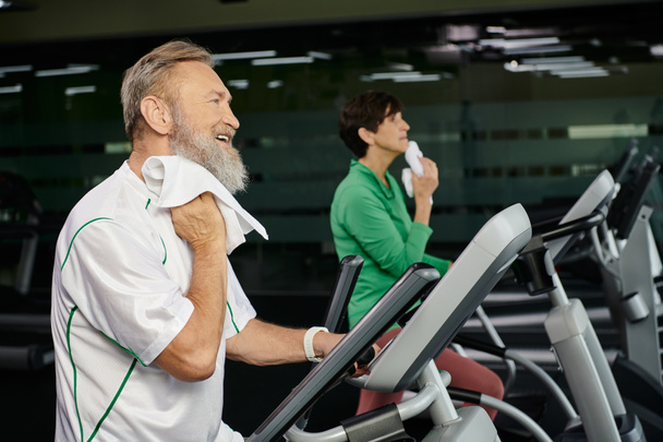 homme âgé et barbu essuyant la sueur avec une serviette, femme floue sur le fond, faisant de l'exercice dans la salle de gym - Photo, image