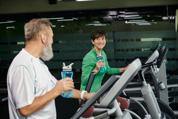 веселая женщина смотрит на пожилого мужчину, мужа и жену, тренирующихся в спортзале, держащих в руках спортивные бутылки - Фото, изображение