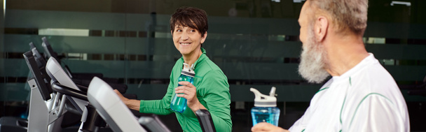 glückliche Frau blickt älteren Mann, Mann und Frau im Fitnessstudio an, hält Sportflaschen, Banner - Foto, Bild