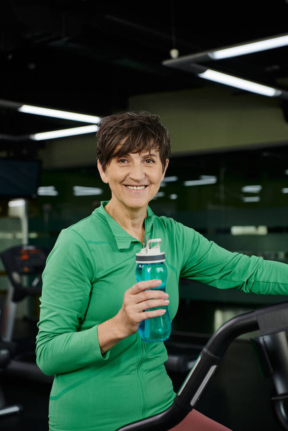 счастливая пожилая женщина держит спортивную бутылку с водой и смотрит на камеру, спорт, тренажерный зал, тренировки - Фото, изображение