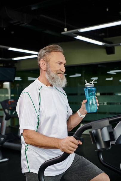 счастливый пожилой и бородатый мужчина, держа в руках спортивную бутылку с водой и отводя взгляд, спорт, тренажерный зал, тренировка - Фото, изображение