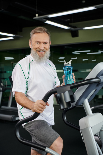 Yaşlı ve sakallı adam elinde su şişesiyle kameraya, spora, spor salonuna bakıp gülümsüyor. - Fotoğraf, Görsel