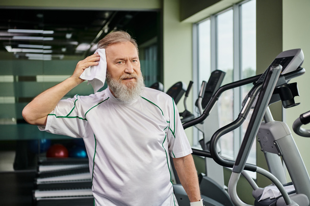 Spor salonunda spor yaptıktan sonra terini havluyla silen sakallı yaşlı adam kameraya ve spora bakıyor. - Fotoğraf, Görsel
