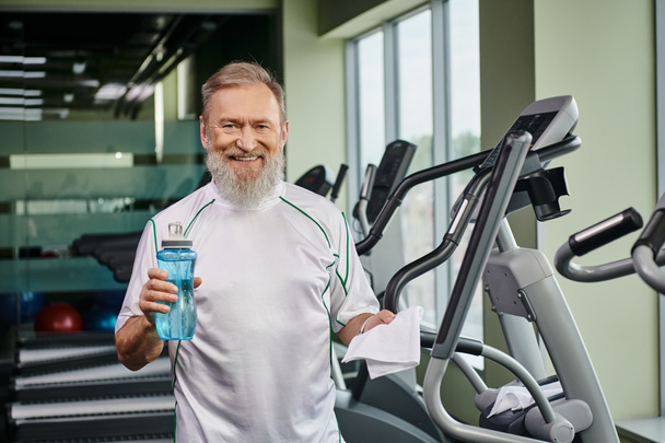 χαρούμενος ηλικιωμένος άνδρας κρατώντας αθλητικό μπουκάλι με νερό και χαμογελώντας μετά την άσκηση στον αθλητισμό γυμναστήριο - Φωτογραφία, εικόνα