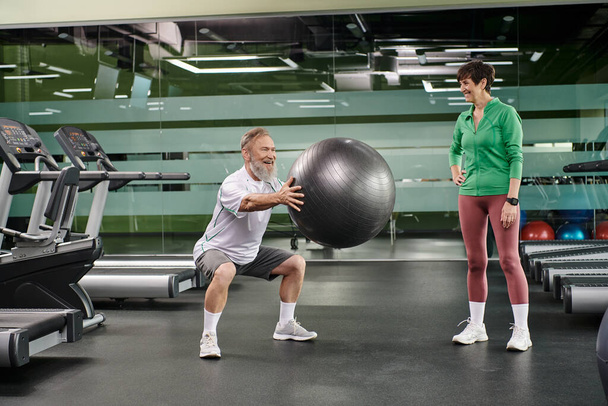aufgeregter älterer Mann macht Kniebeugen mit Fitnessball in der Nähe von Frau, Mann und Frau in der Turnhalle, Paar, Sport - Foto, Bild