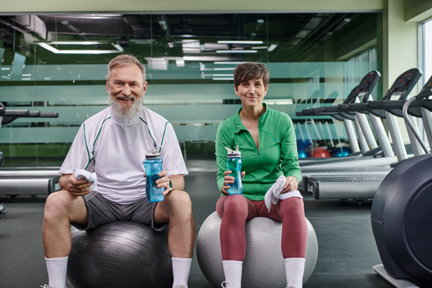 спортивная пожилая пара, счастливый мужчина и женщина сидят на фитнес-мячах, держа бутылки с водой - Фото, изображение