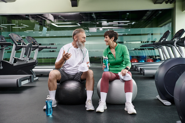спортивная пожилая пара, весёлый мужчина и женщина сидят на фитнес-мячах, держа бутылки с водой - Фото, изображение