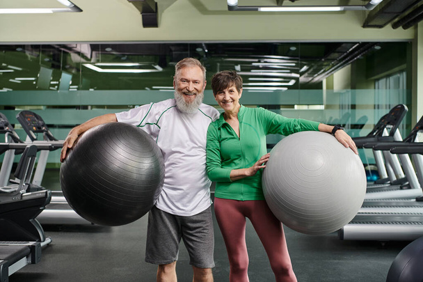 спортивная пожилая пара, веселый мужчина и женщина, держащие фитнес-мячи, активные пожилые люди в тренажерном зале - Фото, изображение