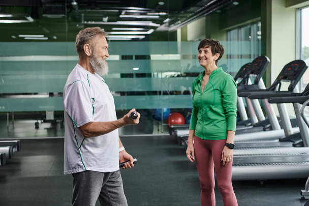 счастливая женщина смотрит на бородатого мужа, тренирующегося с гантелями в тренажерном зале, активных пожилых людей, образ жизни - Фото, изображение