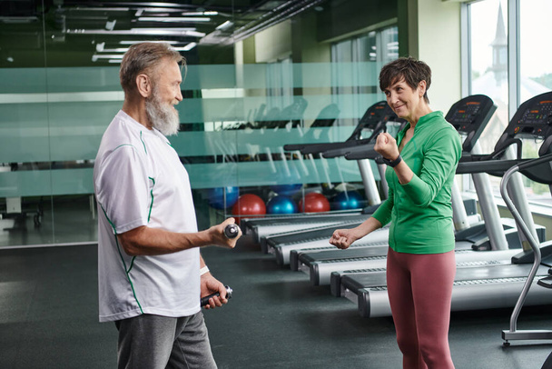 szczęśliwa kobieta patrząc na brodatego mężczyznę ćwiczącego z hantlami na siłowni, aktywnych seniorów, styl życia - Zdjęcie, obraz