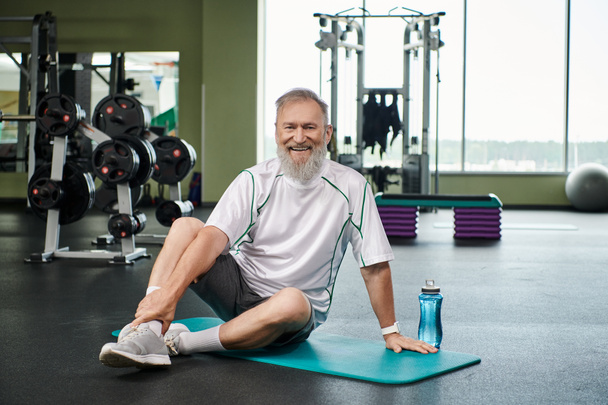 cheerful elderly man with beard sitting on fitness mat near sports bottle with water, active senior - Photo, Image