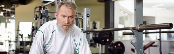 tired elderly man with beard looking at camera after workout, exercise machine in gym, banner - Photo, Image