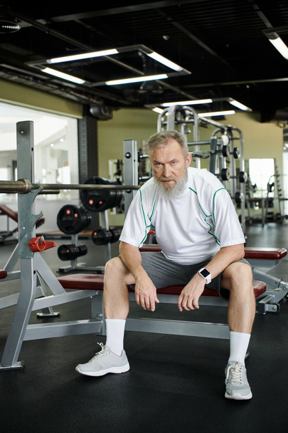 zmęczony staruszek z brodą patrzący w kamerę po treningu, siedzący na trenażerze w siłowni - Zdjęcie, obraz
