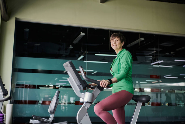 feliz anciana con pelo corto haciendo ejercicio en bicicleta estática en el gimnasio, activa, motivación - Foto, Imagen