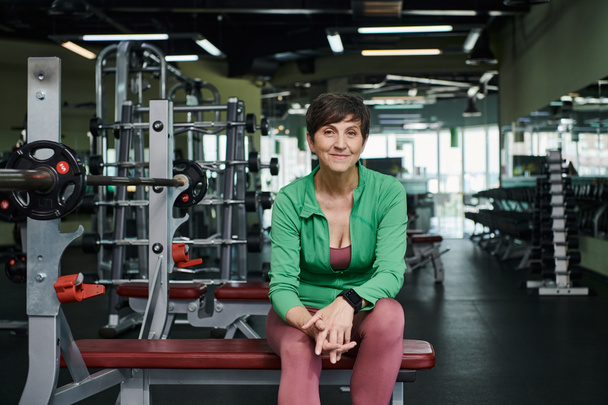feliz anciana sonriendo y sentada cerca de la barra, placas de peso, entrenamiento en el gimnasio, motivación - Foto, Imagen