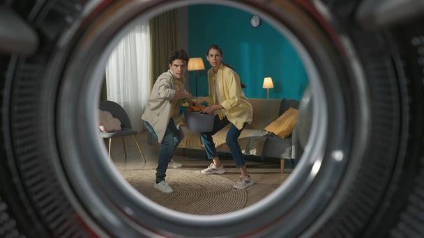 Young couple doing laundry. Pair funnily throwing clothes inside the empty drum. Couple having fun doing home chores. View from inside the empty washing machine. - Fotografie, Obrázek