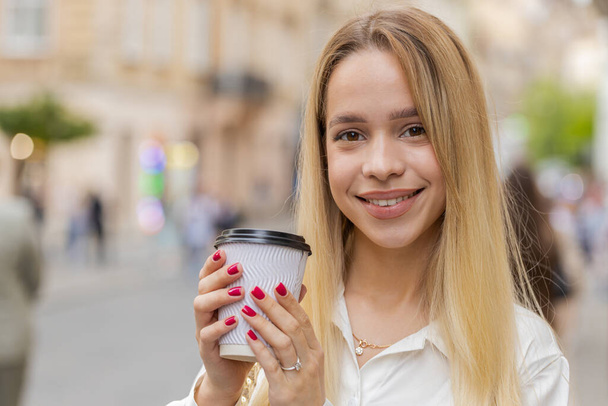 朝のコーヒーホットドリンクを楽しみ、屋外で笑顔の幸せなブロンドの若い女性。リラックスして休憩。10代の少女都市部の街を歩くと、移動するコーヒーを飲む。外の町の生活 - 写真・画像