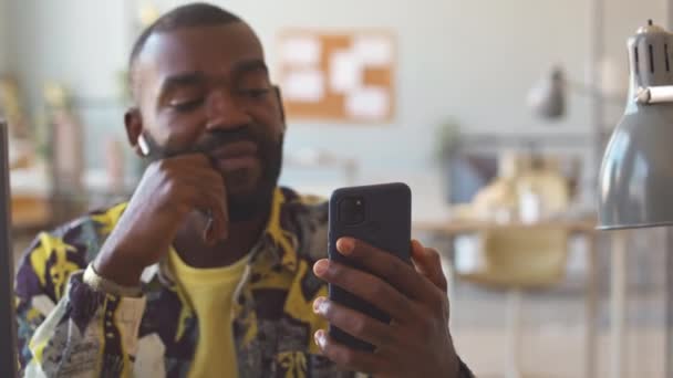 Joven hombre negro en colorido camisa impresa videollamadas con colega en el teléfono inteligente a través de auriculares inalámbricos sentados en su lugar de trabajo en la oficina - Imágenes, Vídeo