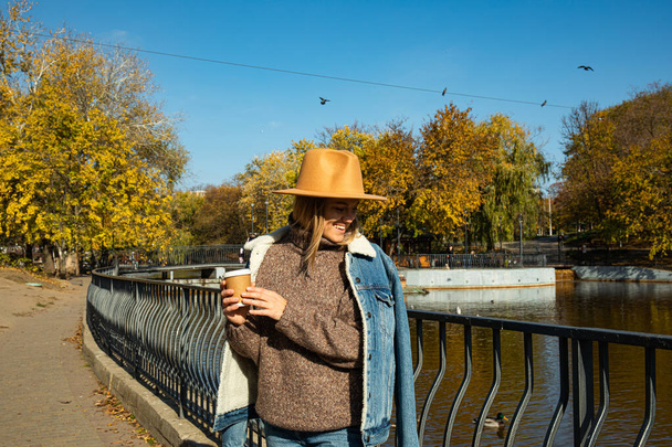 Ein schönes Mädchen mit Hut spaziert mit Kaffee in einem herbstlichen Park - Foto, Bild