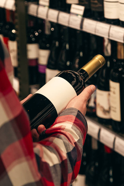 Un hombre sosteniendo una botella de vino en una tienda de licores, eligiendo el vino adecuado de todas las variaciones de botellas de vino en los estantes. - Foto, imagen