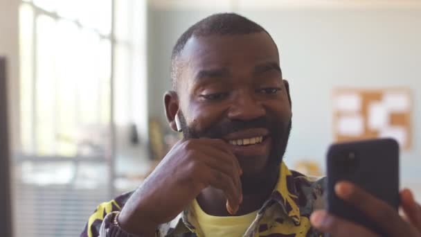 Αφροαμερικανός σε ασύρματα ακουστικά βίντεο κουβεντιάζοντας στο smartphone με συνάδελφο στο γραφείο - Πλάνα, βίντεο