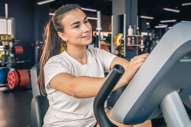 A beautiful athletic young brunette woman in sportswear in the gym is engaged on a bike, healthy lifestyle. - Photo, Image