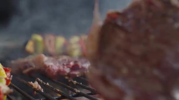 Detailní záběr otáčení šťavnaté masové steaky na BBQ s kovovými kleštěmi, zatímco grilování je s veggie špejlemi - Záběry, video