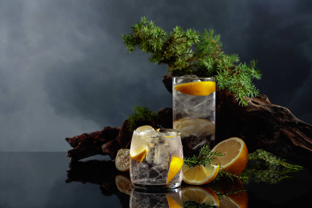 Cocktail Gin Tonic mit Eis und Zitrone auf schwarzem, reflektierendem Hintergrund. Eisgetränk mit alten Haken und Wacholderzweigen. - Foto, Bild