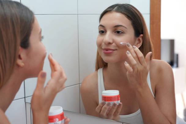 Beauty woman holds container with cosmetic cream and applies on her cheek at home - Photo, Image