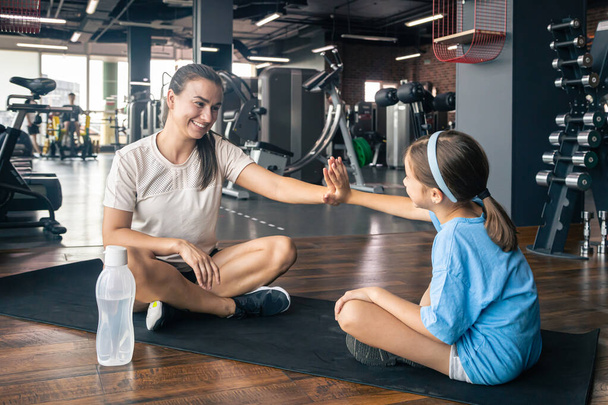 Side view of strong mother and daughter sitting on yoga mat and giving high five in gym. - Photo, Image