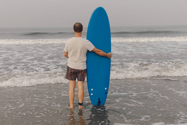 Οπίσθια όψη ώριμου άνδρα με γκρι t-shirt και ριγέ σορτς που στέκεται στο νερό στη θάλασσα και κρατά την μπλε μακριά σανίδα του σερφ κοντά. Surfer στροφή προς κυματίζει θάλασσα και κοιτάζοντας μπροστά στο θαλασσογραφία. - Φωτογραφία, εικόνα