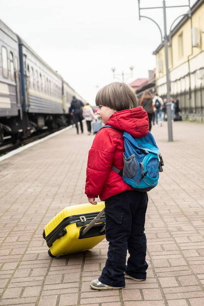  μικρό απρόσωπο αγοράκι στέκεται στην αποβάθρα κοντά στο τρένο και κρατά μια κίτρινη βαλίτσα στα χέρια του. Έννοια μετακίνησης αμαξοστοιχίας - Φωτογραφία, εικόνα