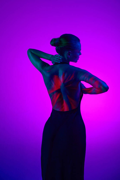Back view. Tender relaxing woman in digital neon filter lights showing red flower on back over gradient purple background. Digital art, fashion, futurism, beauty, concept - Zdjęcie, obraz